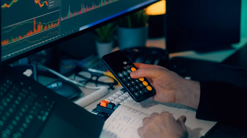 Calculator on a desk beside a computer showing charts